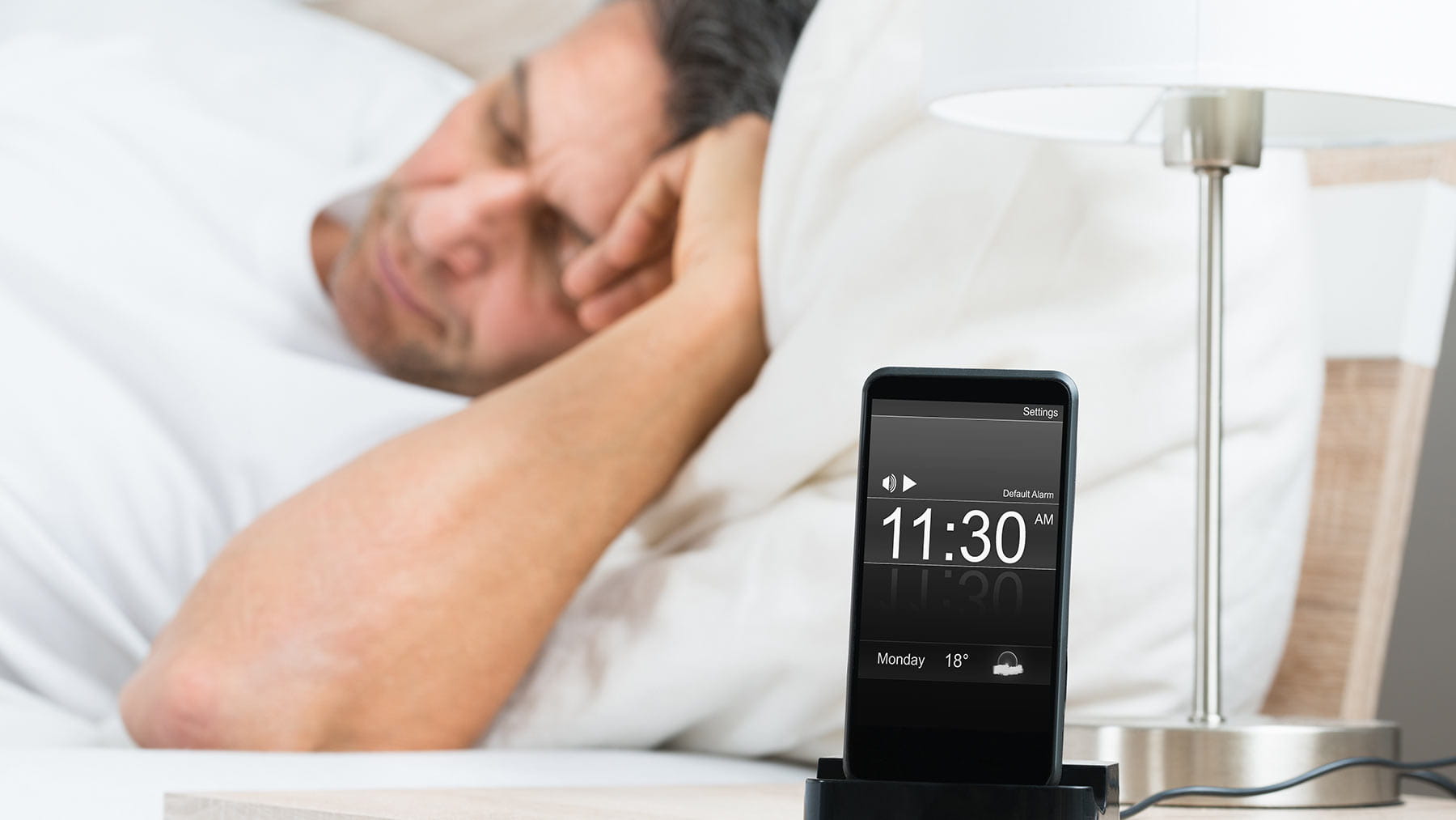 man sleeping in bed with alarm clock set for 11:30 a.m.