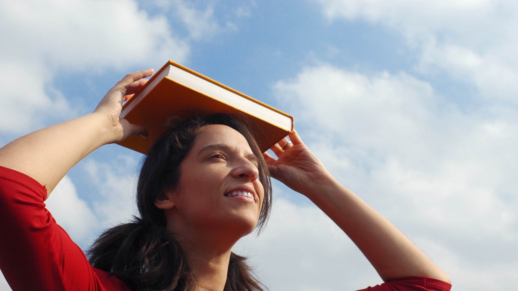 woman balancing book on her head with blue sky and white clouds in background
