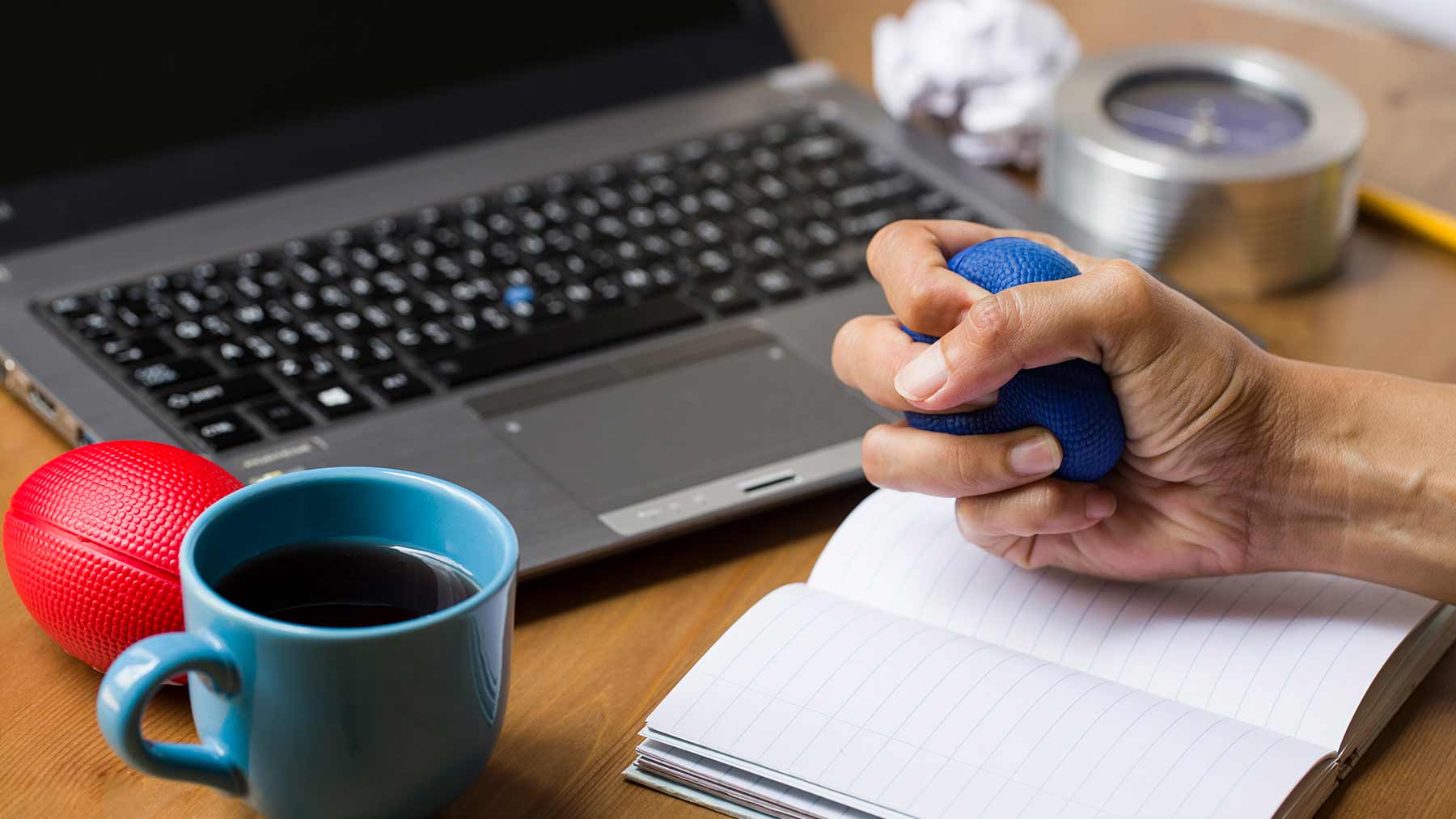 a hand squeezing a stress ball near a cup of coffee and laptop computer
