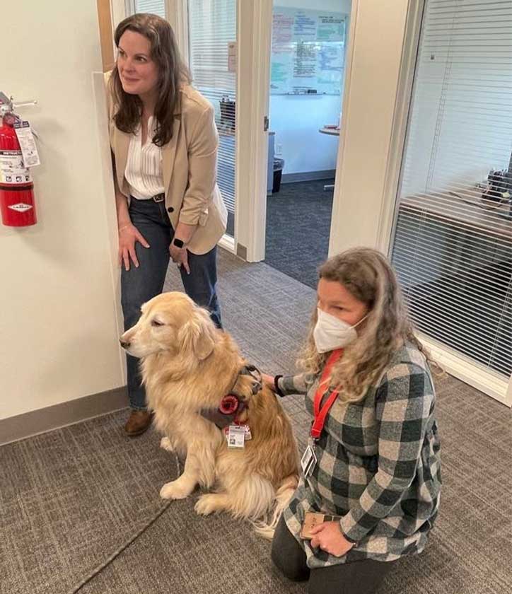 One woman stands while another kneels next to a golden retriever.