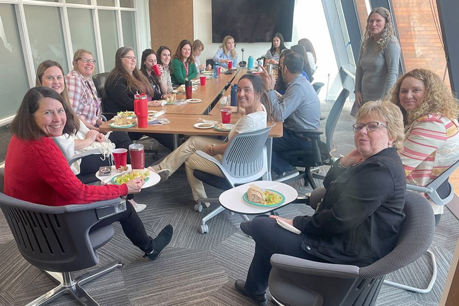 A group of nearly 20 CATALYST faculty and staff members smile toward the camera while enjoying a lunch together at a large table in a meeting room.
