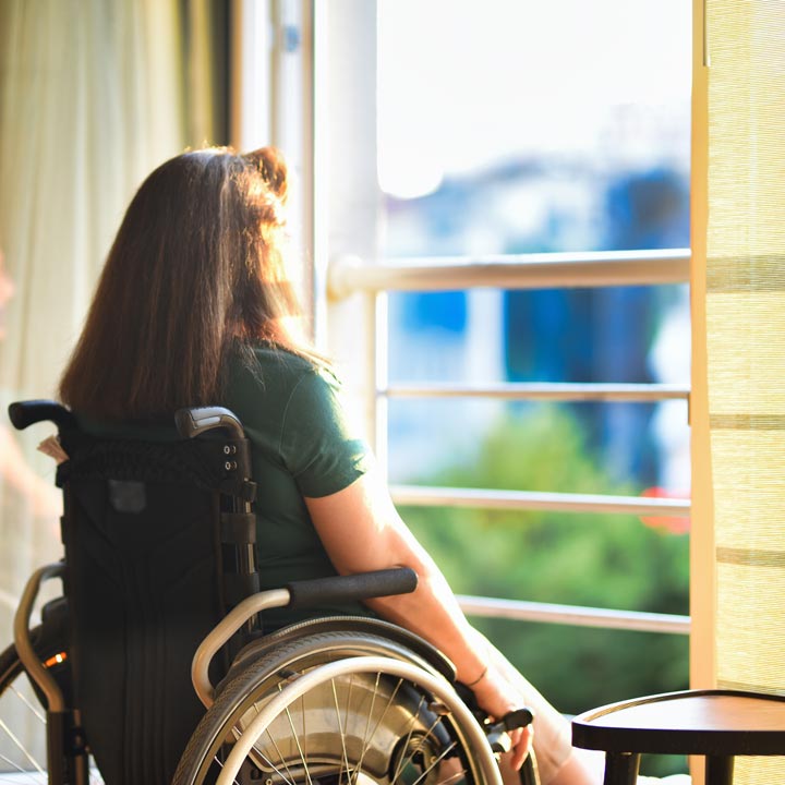 Woman in wheelchair in front of open window in her apartment watching sunset