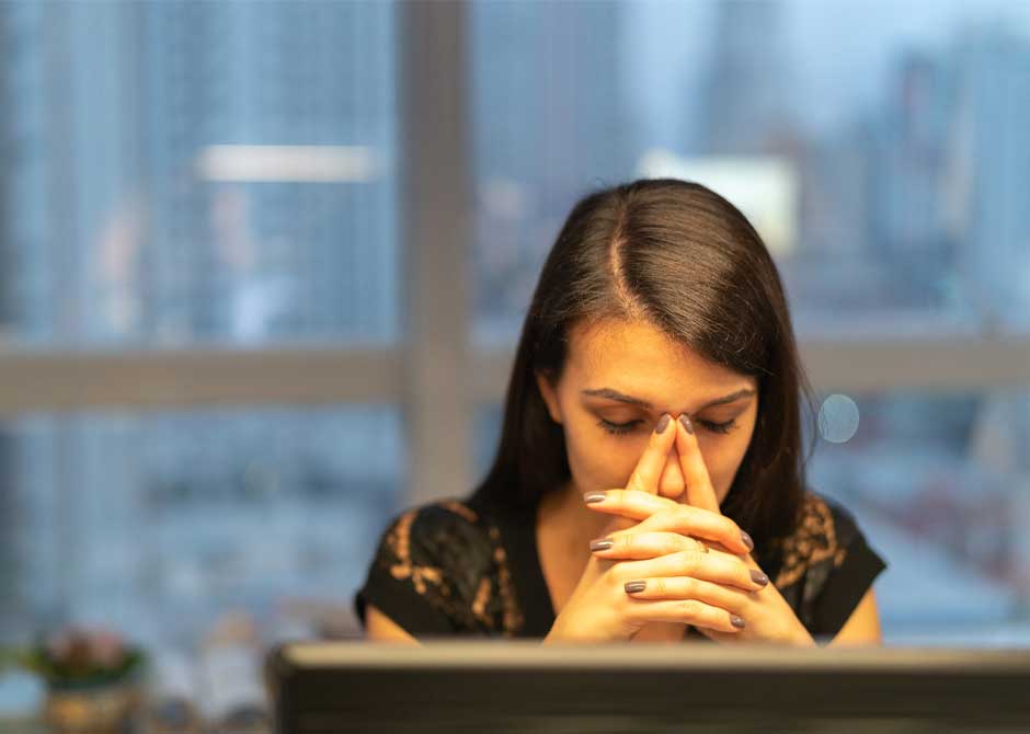 Woman sitting in front of computer monitor with eyes closed and head resting on folded hands