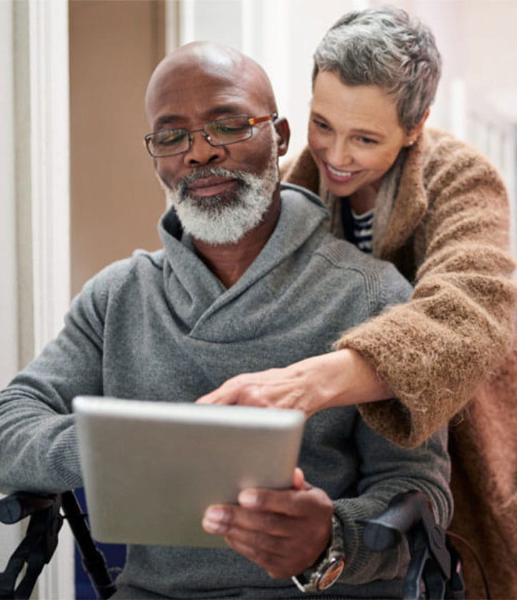 Woman standing over a man, resting her forearms on his shoulder, both looking at a tablet. 