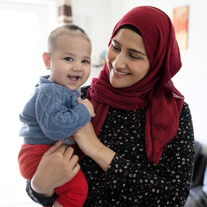 Partial front view of early 30s woman in hijab and tunic smiling at 5 month old boy in her arms as he looks at camera and laughs.