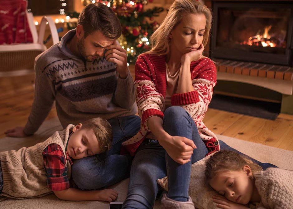 Parents appear annoyed at each other while two kids sleep at their feed in living room with holiday tree.